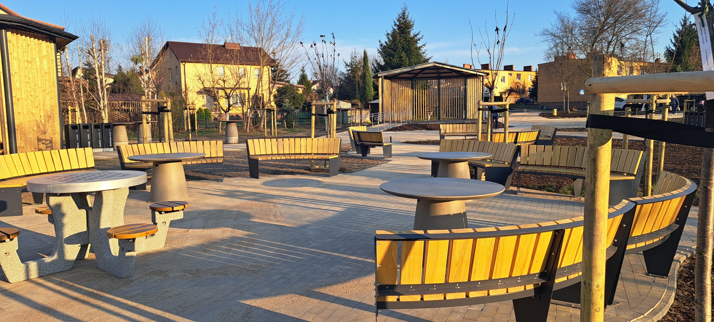 Plac pokryty kostką brukową, stoją na nim ławki i betonowe chodniki. Na końcu placu stoi mini scena. Na drugim planie widoczne budynki mieszkalne